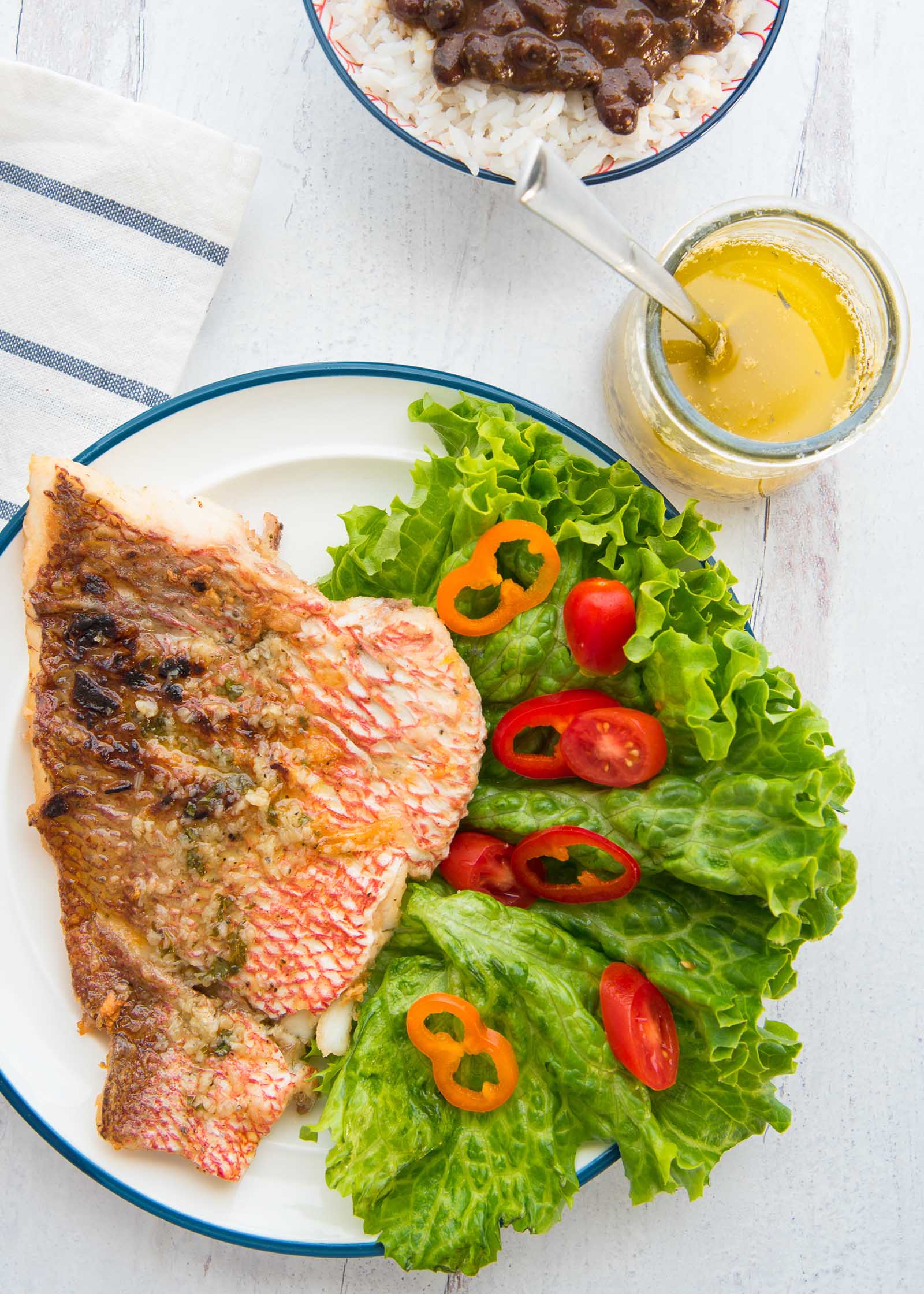 Red snapper with garlic sauce is on a white plate with a blue stripe around the edge. The snapper is crispy and takes up half of the plate. A green leafy salad with red and orange peppers is on the other side of the plate. A jar of garlic sauce is to the upper right of the salad. A bowl of rice and beans with cilantro is to the upper left of the jar of sauce. A striped napkin are to the left of the snapper.