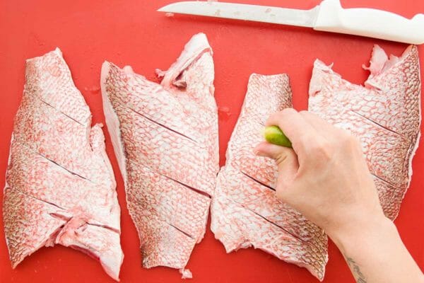 Four skin on fillets of red snapper are on a red cutting board. The skin has been sliced horizontally and lime is being squeezed over top.