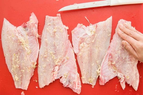 Four raw fillets of red snapper is being coated by hand with a garlic marinade. They sit on a red cutting board and a knife is laid above the fillets.