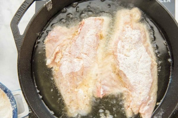 Raw red snapper fillets are side by side in a cast iron skillet. They are coated in flour and frying in oil in a cast iron skillet.