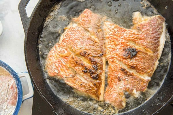 Red snapper with garlic sauce frying in a cast iron skillet. Two fillets are side by side in the pan and liquid is simmering in the pan.