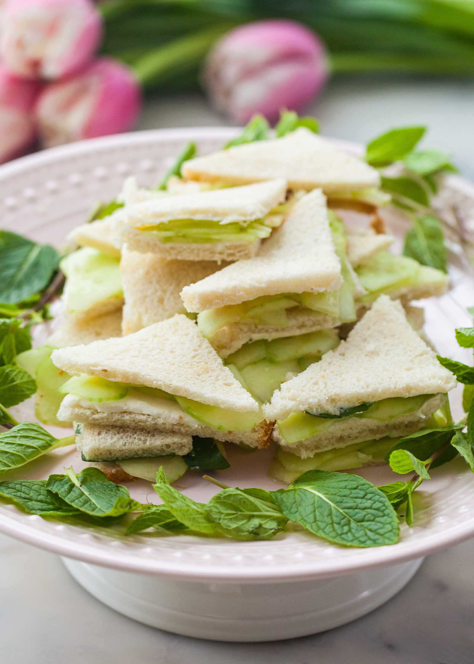 Side view of cucumber finger sandwiches stacked on a cake plate. The sandwiches are cut into triangles and slices of cucumber are visible layered inside each one.