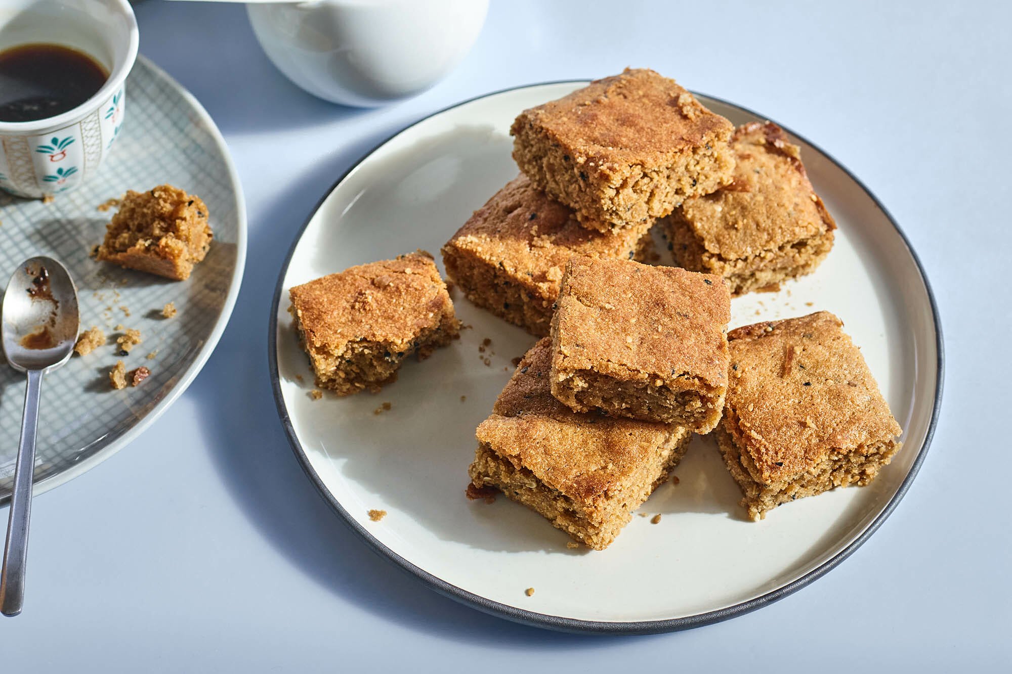 White plate with a stack of square cute chewy tahini blondies on it against a bluish grey background. A cup of coffee with a partially eaten blondie is nearby.