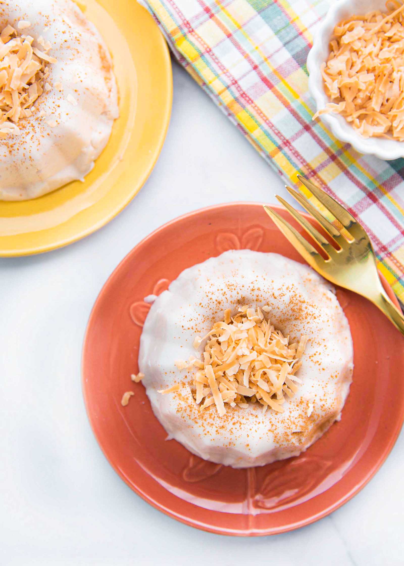 Vertical photo of two plates with coconut pudding with toasted, shredded coconut mounded inside the bundt shape. The coconut pudding is coated in ground cinnamon. The lower plate is coral and a gold fork is to the left of it, the plate in the upper left is light yellow. A small container of toasted shredded coconut is to the right of the coral plate. A striped dish cloth is underneath the coconut.