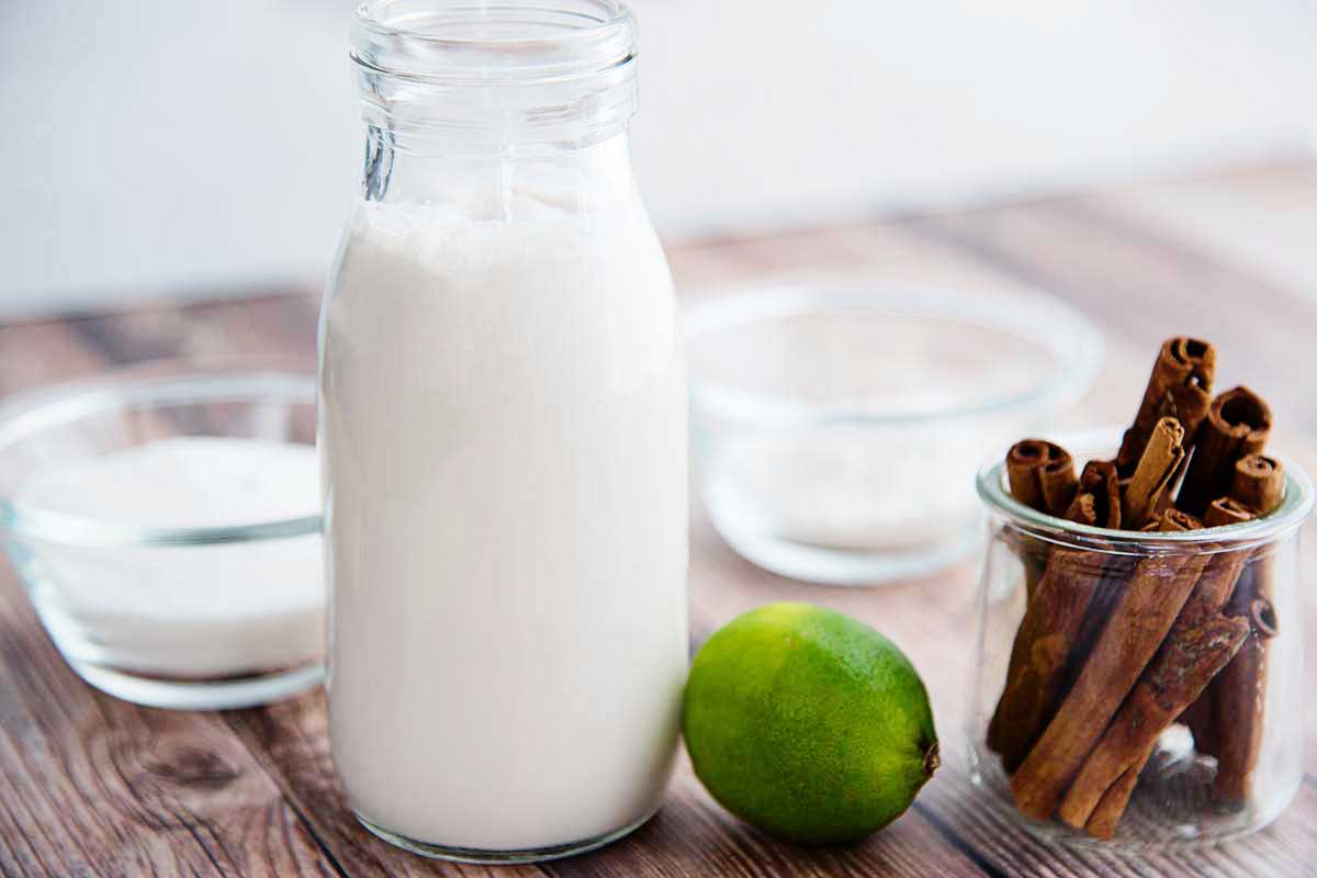 Side view of a glass milk jar with coconut milk insdie. A whole lime and a glass jar of cinnamon sticks are to the right of the milk. Behind the milk are two small glass containters with white ingredients inside.