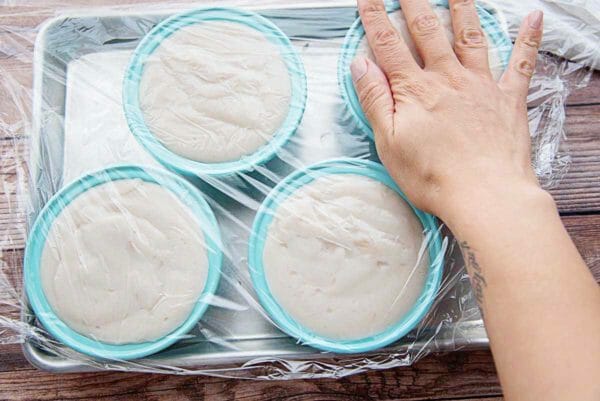 Four teal molds are filled with Puerto Rican Panna Cotta and set on a baking sheet. Plastic wrap covers the molds and a hand is pressing down on the top right mold.