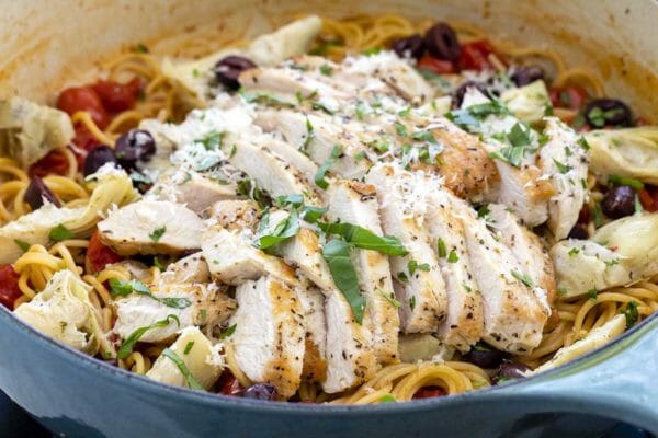 One pot chicken pasta dinner with sliced chicken laid on top of a pot of pasta, olives, tomatoes and basil.