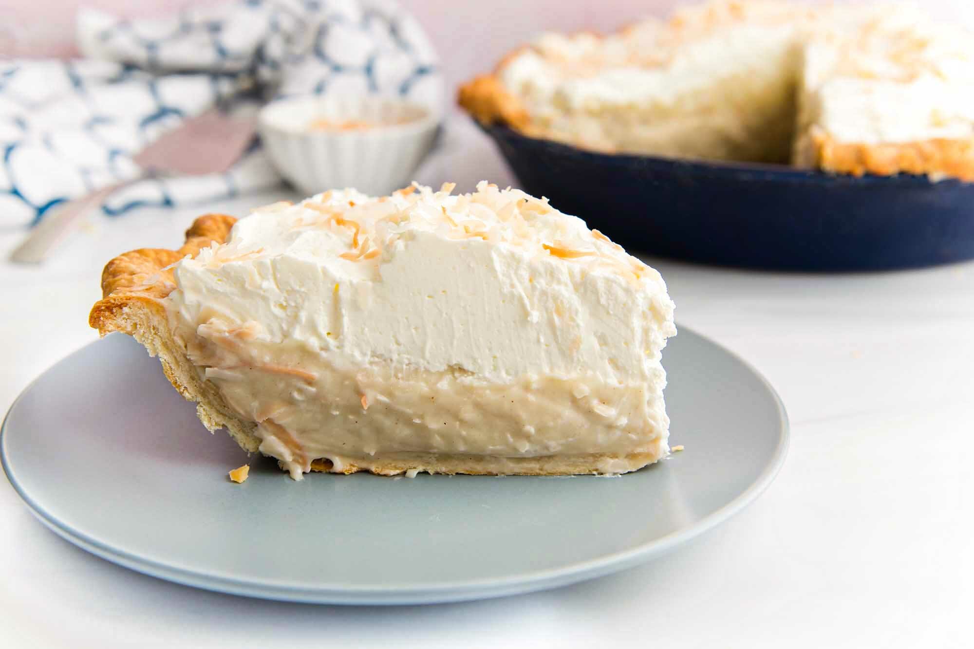 Side view of toasted coconut cream pie with a crisp crust, coconut filling and topped with a thick layer of whipping cream. The rest of the pie is visible behind the plate.
