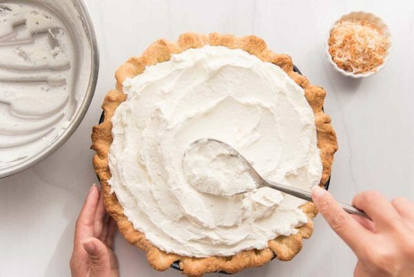 A spoon spreads whipped cream over the top of the best coconut cream pie.