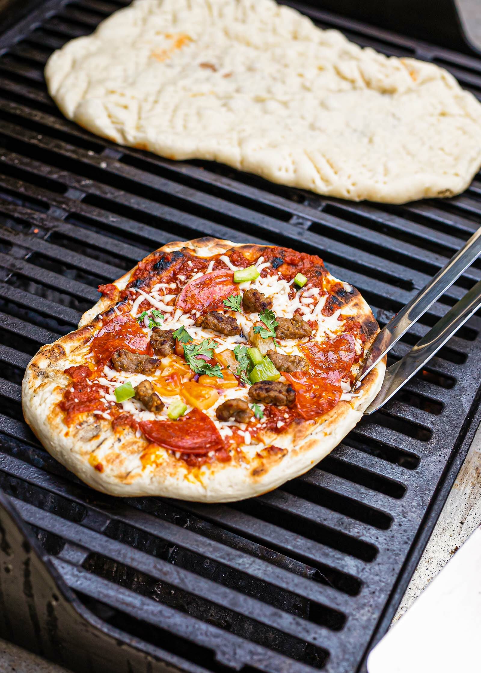 A pizza is on the grill and loaded with toppings and tongs hold it. A rectangular piece of dough is on the other side of the grill to show how to make pizza on the grill.