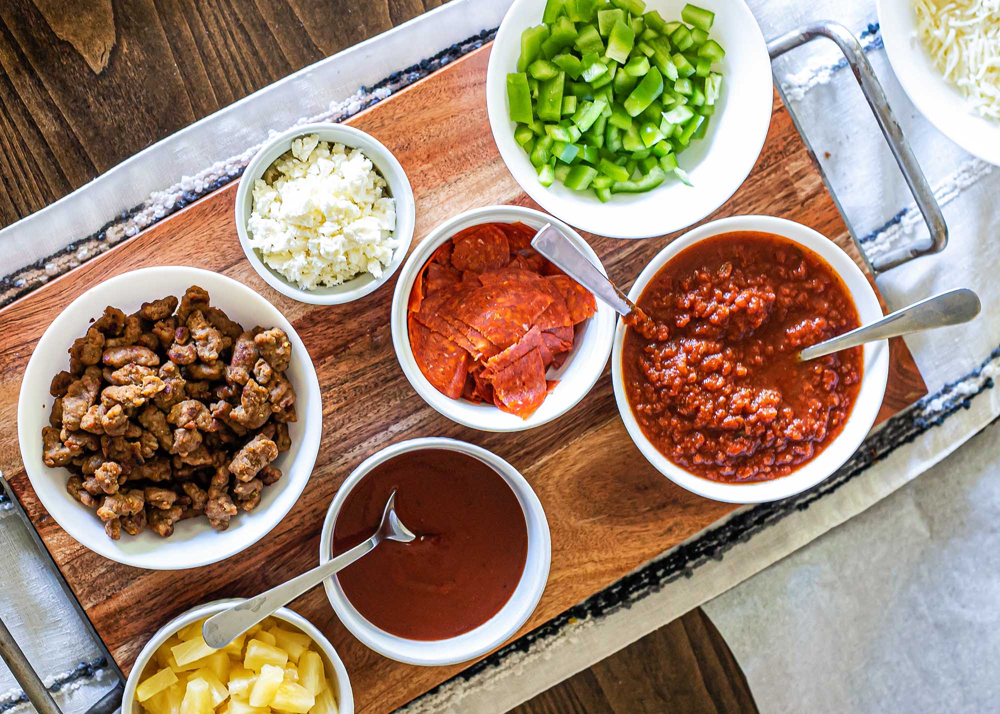 Overhead view of a wooden tray on a table with bowls of pizza toppings.