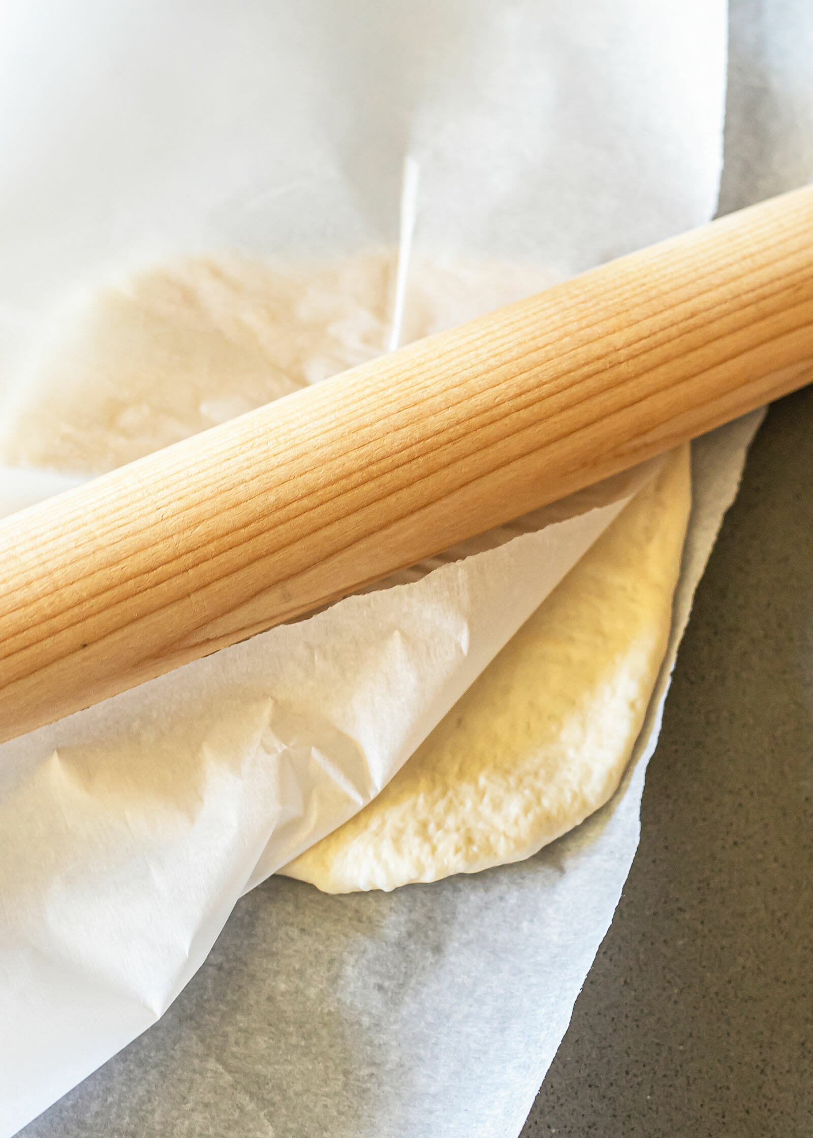 A rolling pin rolls out pizza between pieced of parchment paper.