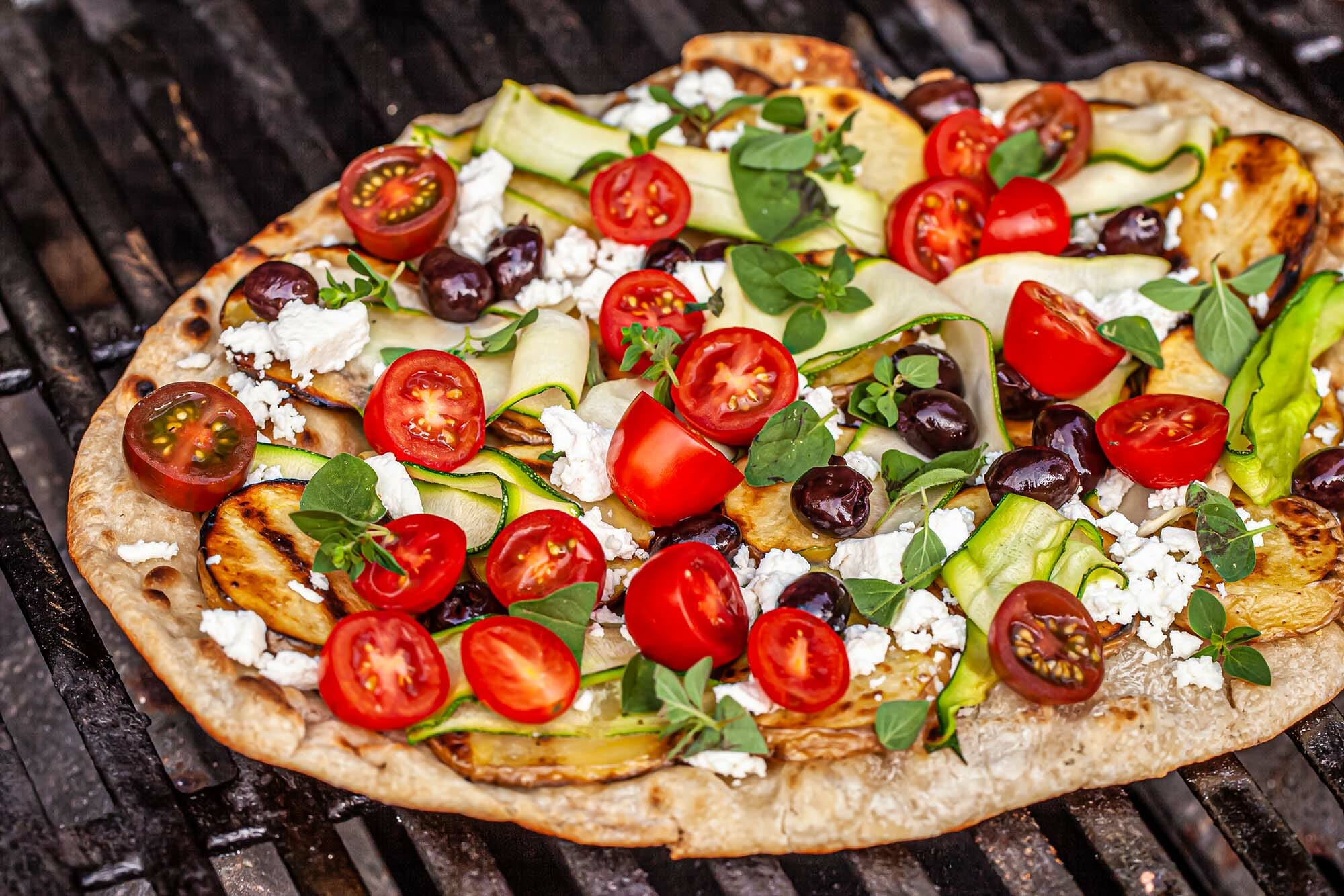 Grilled greek pizza on a grill and topped with halved tomatoes, black olives, cheese, zucchini ribbons and sliced potatoes.