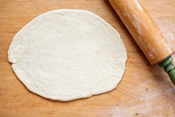 Pizza dough rolled out into a thin circle on a wooden counter top. A wooden rolling pin is to the right.