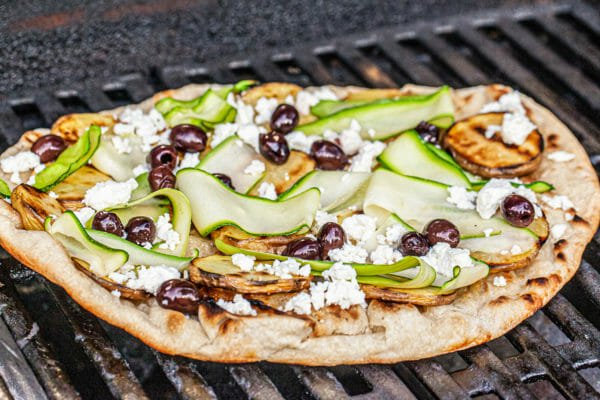 Pizza dough on a grill topped with strips of zucchini, olives and cheese to make a healthy grilled pizza..