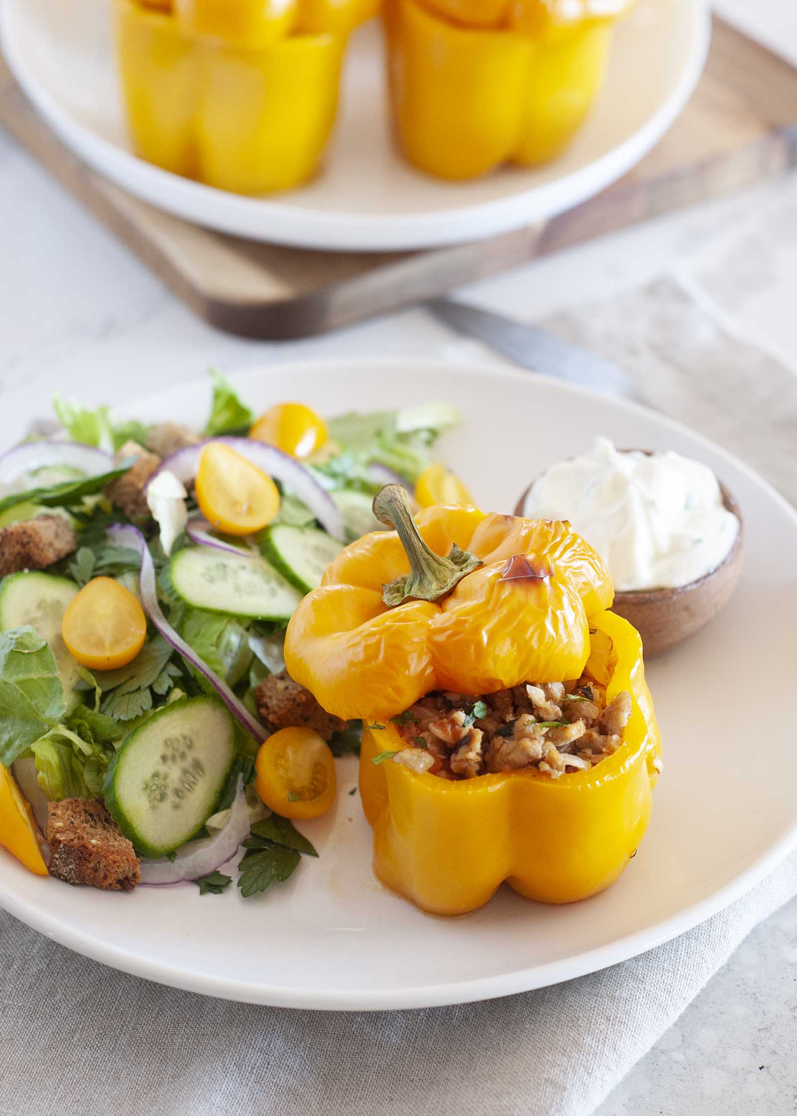 Healthy stuffed pepper with chicken is on a plate with the top askew to reveal the ground chicken filling. A yogurt sauce and garden salad are on the plate as well. Additional peppers are behind the plate.